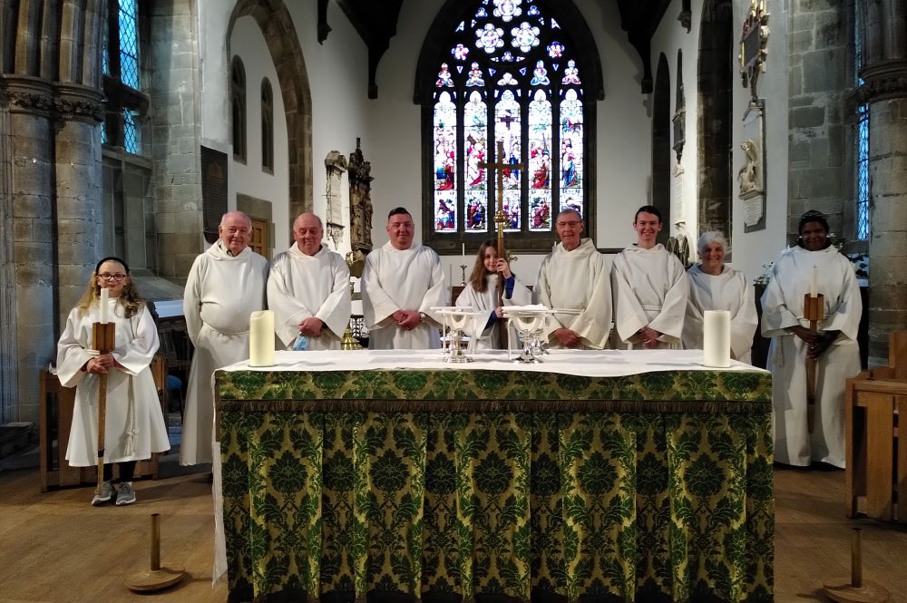 Nine people wearing white serving robes standing behind the altar table. Two are holding processional candles and one a processional cross.